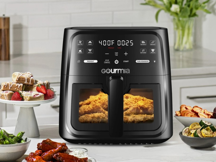 a black Gourmia air fryer with window with food cooking inside, plates of food around it on a kitchen counter