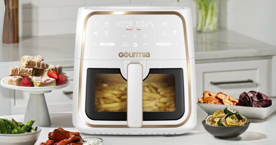 a white and gold Gourmia air fryer with window with food cooking inside, plates of food around it on a kitchen counter