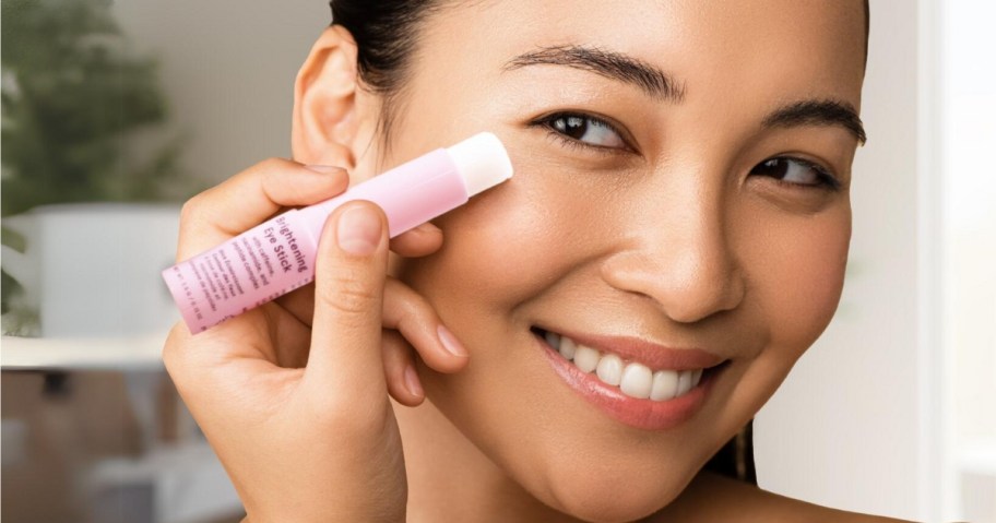 woman holding a pink Under Eye Brightener balm stick, smiling at the camera while applying it under her eyes