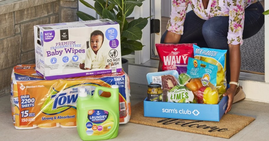 woman picking up a box of groceries in a Sam's Club box from a delivery to her home, large box of diapers, package of paper towels and large laundry detergent next to it