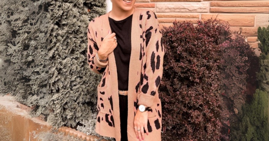 woman standing outside, shrubbery behind her. She is wearing a tan and black animal print long cardigan sweater with a back top and jeans