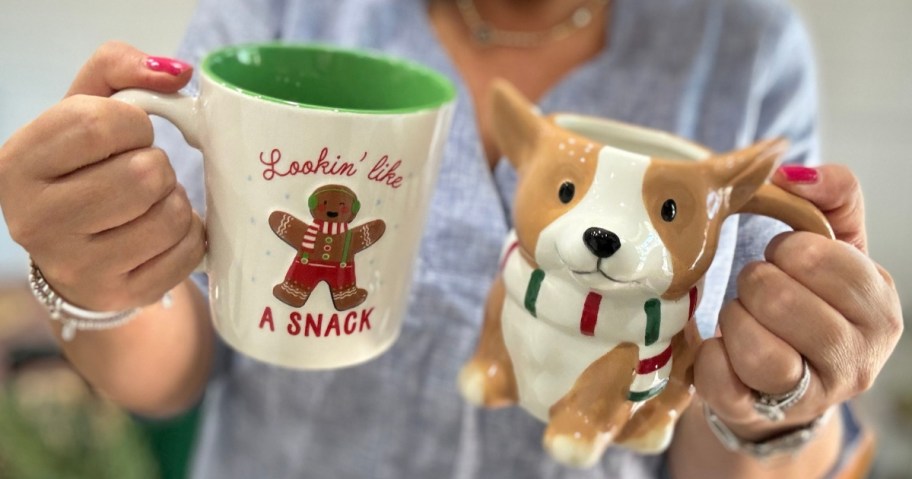 hand holding 2 Christmas mugs, one in white with a gingerbread man that says "Lookin' like a snack" and one shaped like a tan and white Corgi dog with a red, white, and green scarf