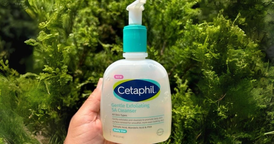 hand holding a clear and blue bottle of Cetaphil Gentle Exfoliating SA Cleanser, greenery in the background