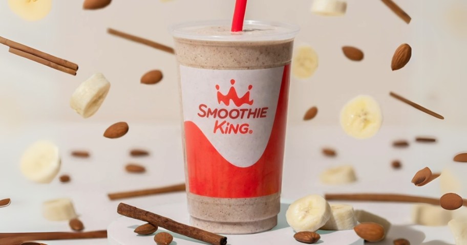 a Smoothie King Power Meal Cinnamon Banana Smoothie in a cup, cinnamon sticks and banana slices all around and behind it