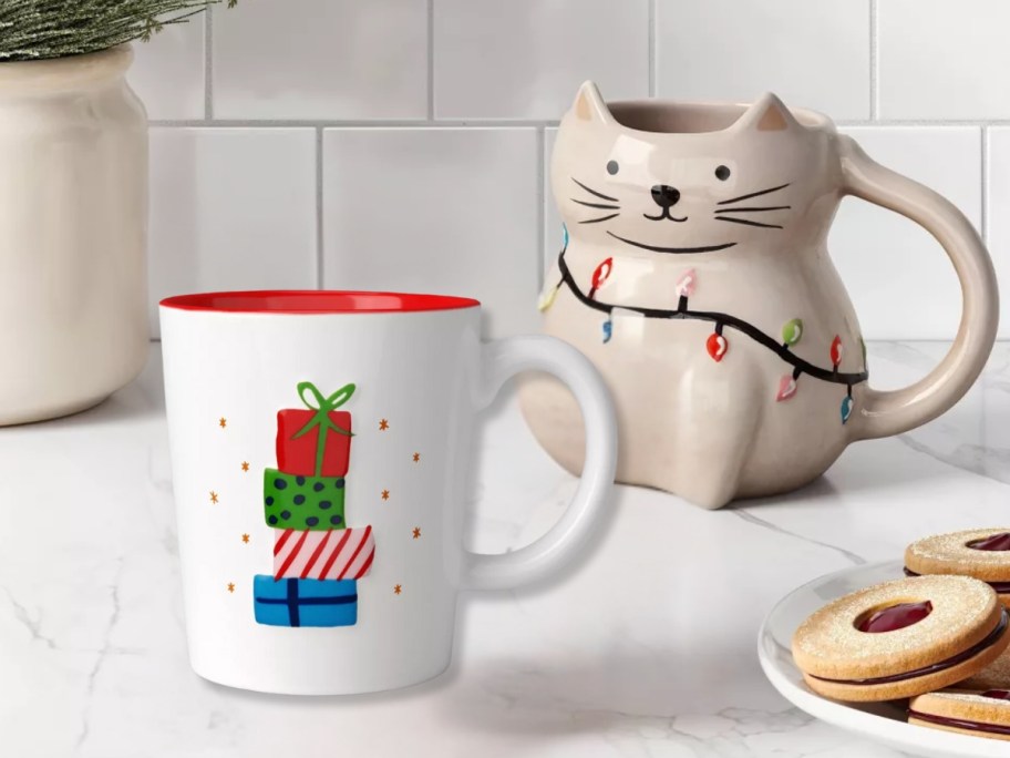 a white coffee mug with Christmas presents and a mug shaped like a tan cat with Christmas lights around it on a kitchen counter