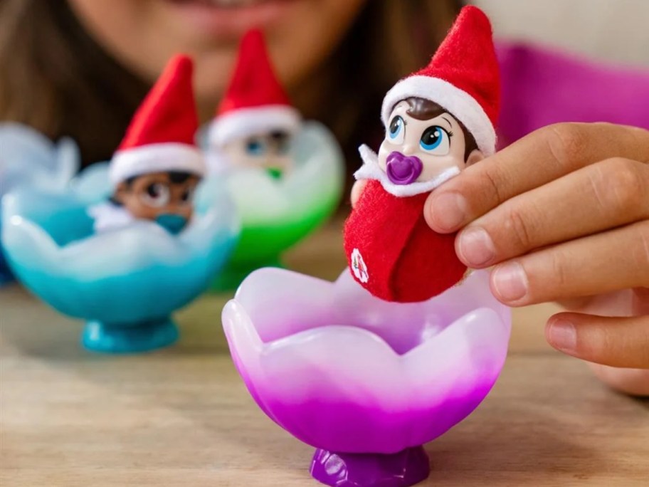 little girl holding a baby Elf on the Shelf Frost Pip above a purple flower cradle, more in the background in other colors