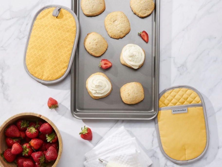two yellow oven mitts shown with a cookie sheet of cookies and a bowl of strawberries