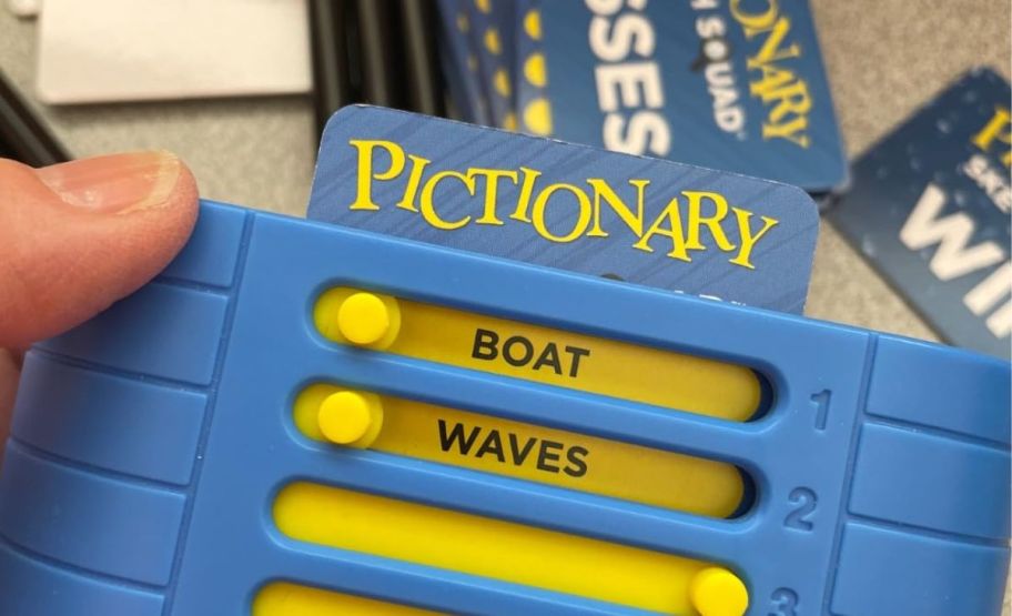 a person's hand holding the Pictionary sketch squad game with cards and markers displayed on a countertop