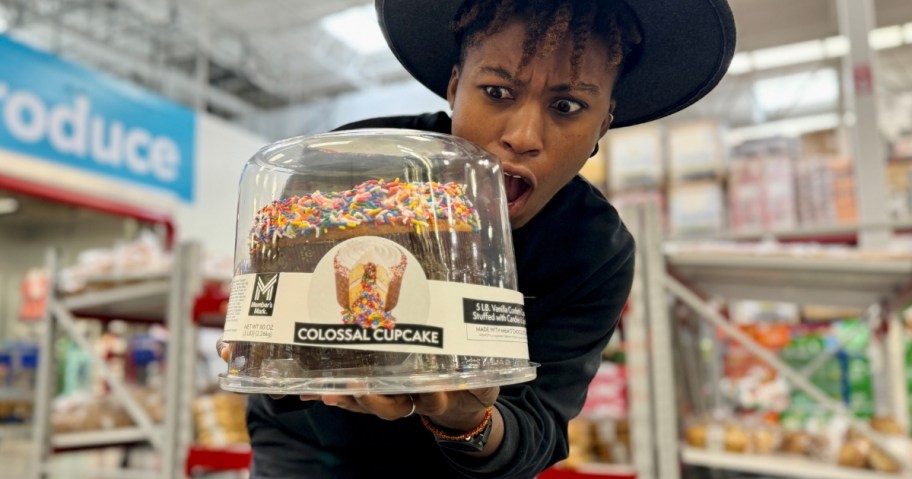 woman with a shocked look on her face looking at a giant cupcake in a plastic container she's holding in her hands