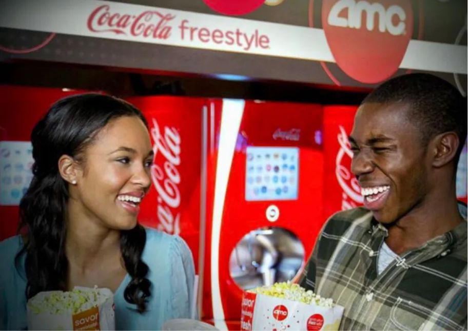 a woman and a man in a movie theatre lobby smiling with popcorn