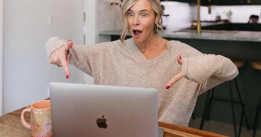 Colling sitting at a table with her laptop in front of her, wearing a tan seater with an excited/shocked look on her face pointing at her laptop