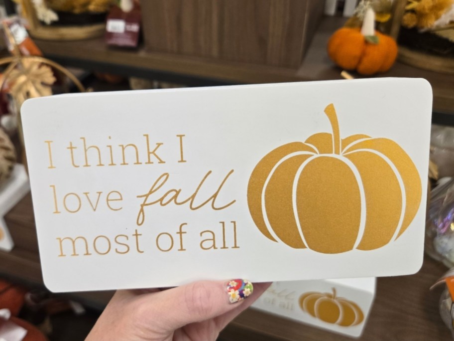 a womans hand holding a sign with words 'I think I love fall most of all' and a pumpkin in gold font and graphics
