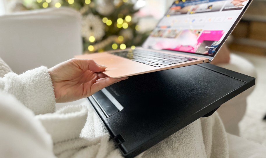 hand holding up laptop with black lapdesk on lap in front of christmas tree