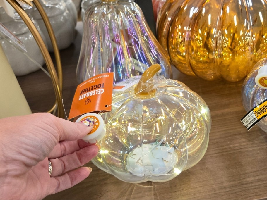 a womans hand turning on the LED lights in a small clear glass pumpkin