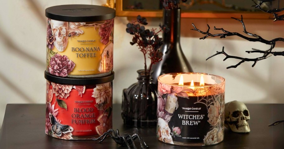 3 Yankee Candle Halloween scent 3-wick jar candles, one lit, 2 stacked with the lids on, on a console table with other Halloween decor