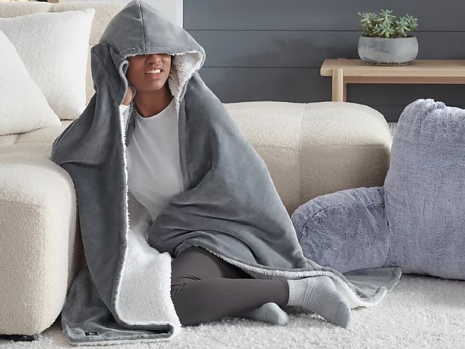 person sitting on the floor by a couch wearing a grey UGG hooded throw