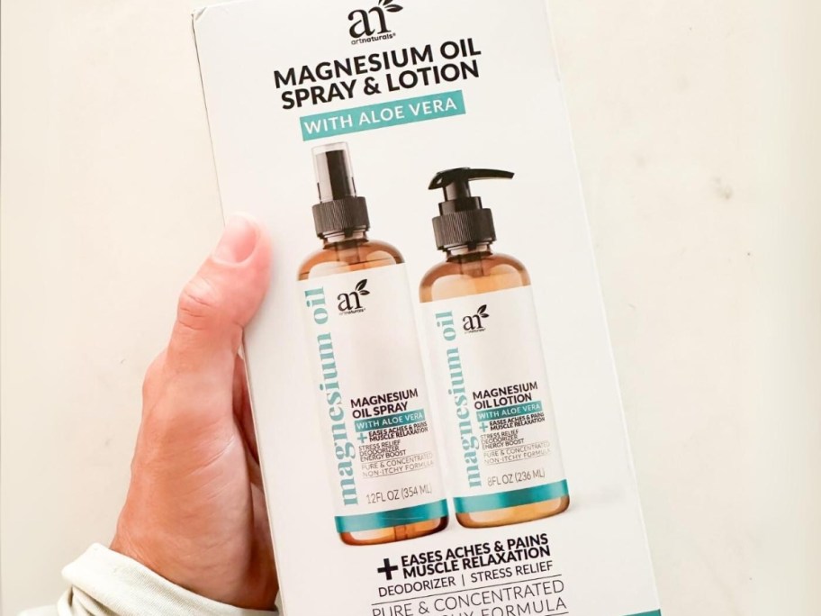 hand holding a white box with 2 brown bottles with white and blue/green labels sitting on a table - one is ArtNaturals Magnesiums Spray and the other is Magnesium Lotion