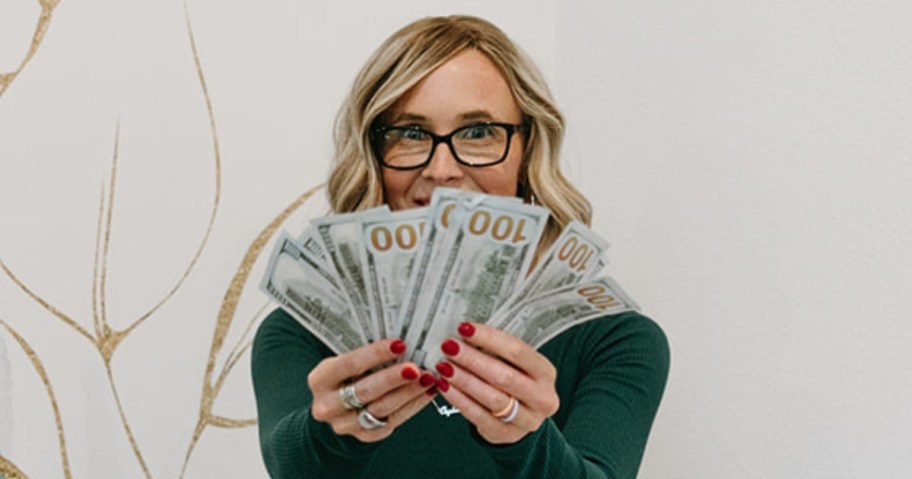 Collin (woman with blond hair and glasses) wearing a green shirt holding up a bunch of $100 bills to the camera