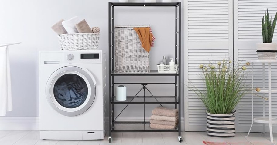 a black adjustable folding metal shelf with wheels in a laundry room next to a washing machine, shelf has laundry hamper, towels, and other laundry items on it