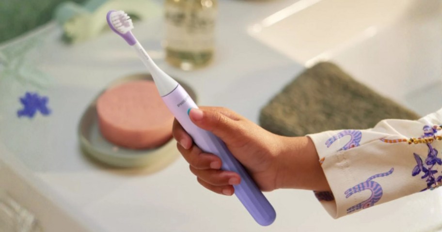 kids hand holding a purple and white electric toothbrush above a bathroom sink