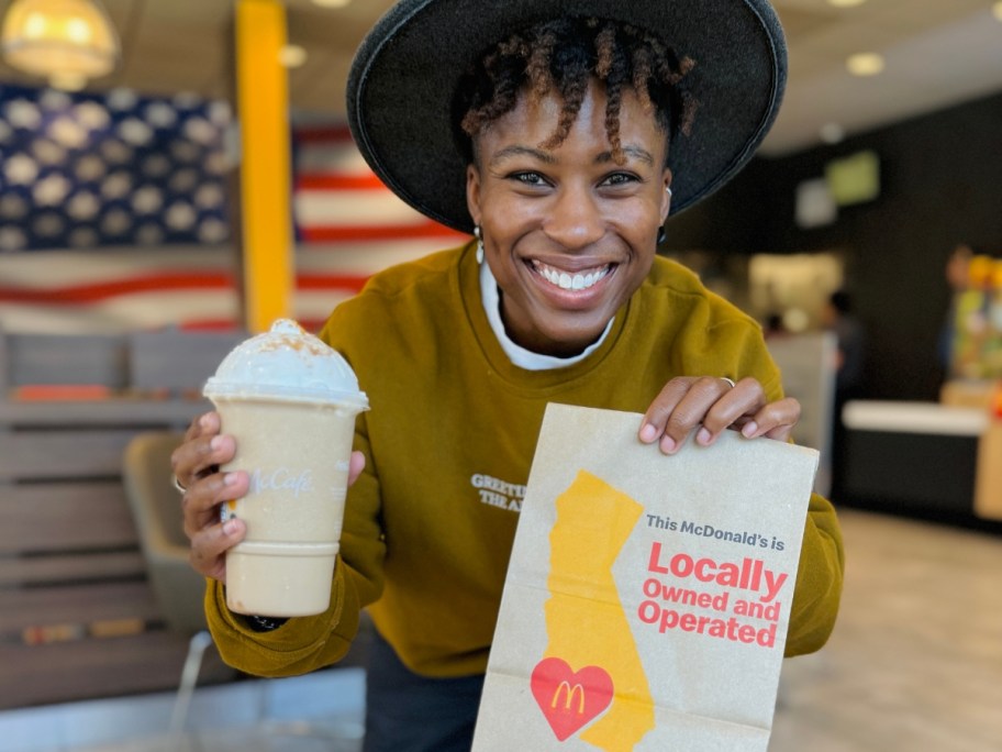 woman smiling at the camera holding a McDonald's cup with a Dulce De Leche Frappe in it and a Mcdonald's paper bag in the other hand