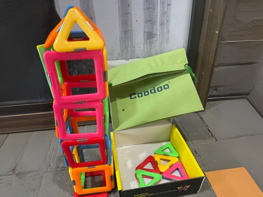 a set of colorful magnetic building tiles shaped like a house, the box they come in and a green storage bag sits to the side