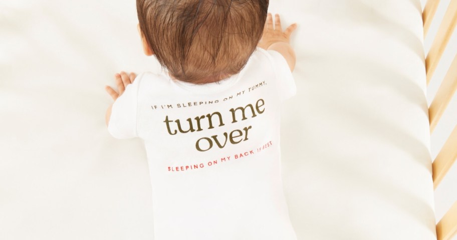 a baby in a crib on it's knees and hands wearing a white short sleeve onesie that says "turn me over"