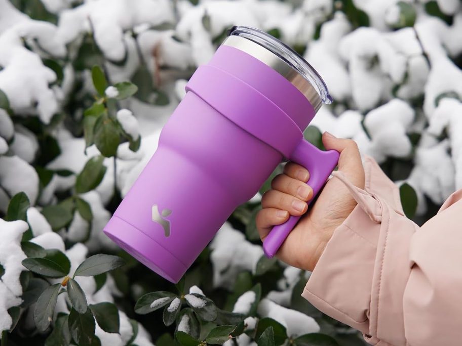 hand holding purple tumbler in front of snow covered bush