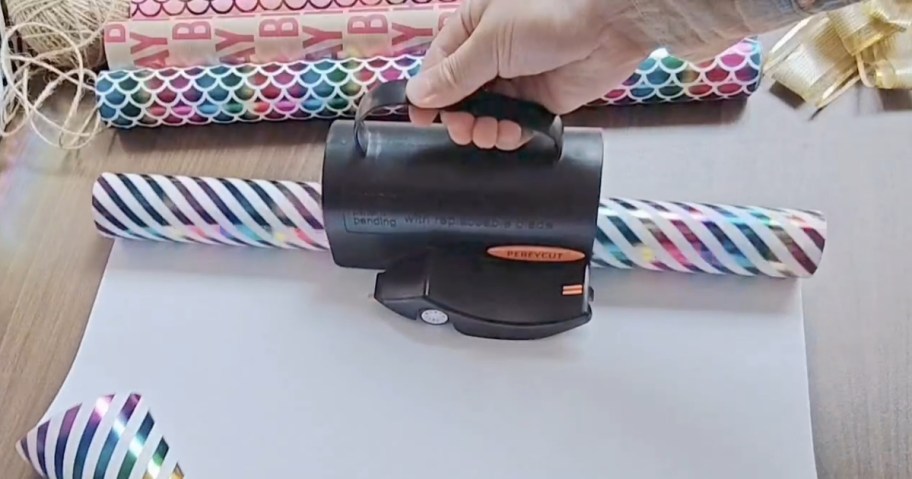 hand on a black and orange wrapping paper cutter cutting through birthday wrapping paper, more wrapping paper behind it