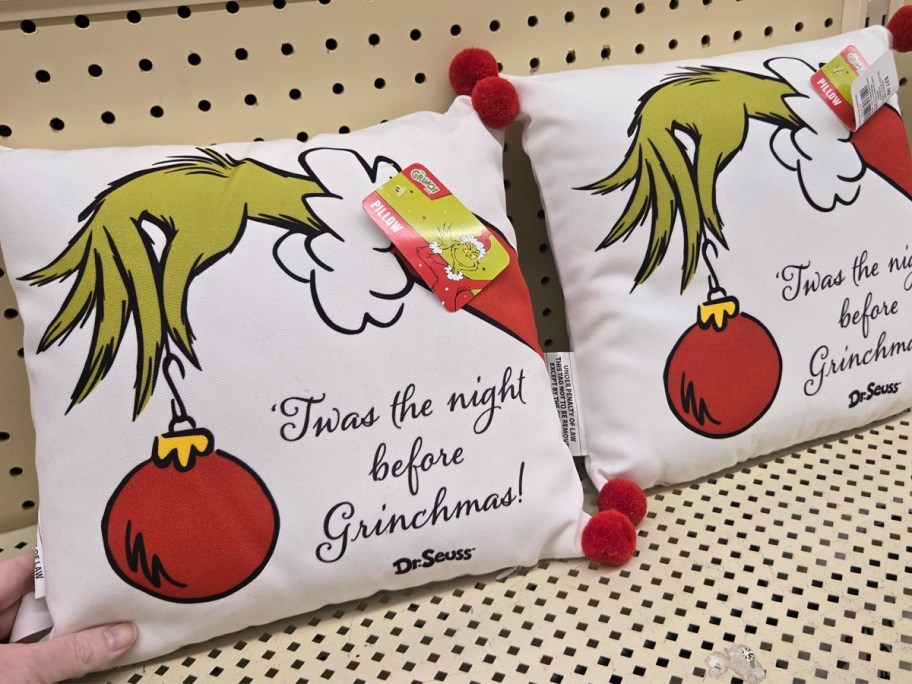 A throw pillow with The Grinch's hand holding a Christmas ornament sitting on a shelf. 