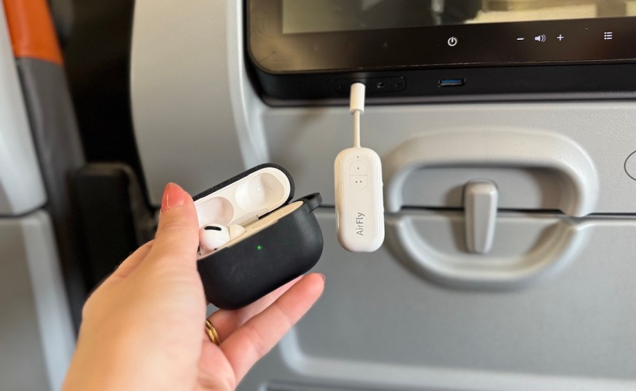 hand holding set of airpoods in case with airfly bluetooth attachment on airplane