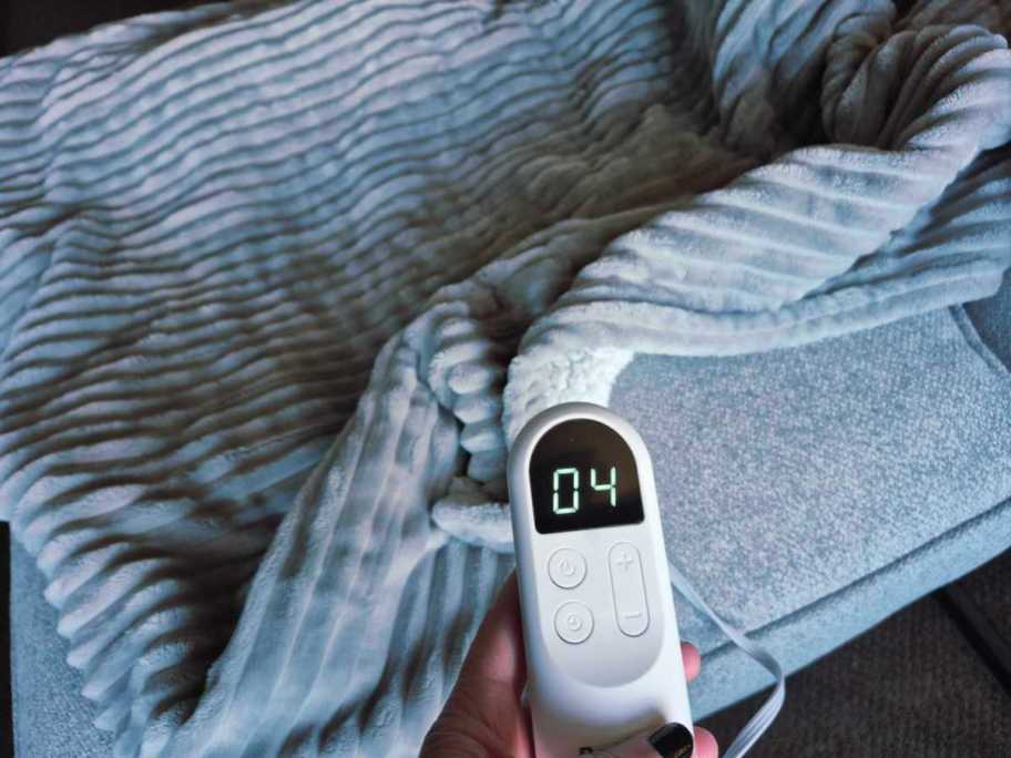 Close-up of hand holding heated blanket remote control with gray ribbed blanket and background