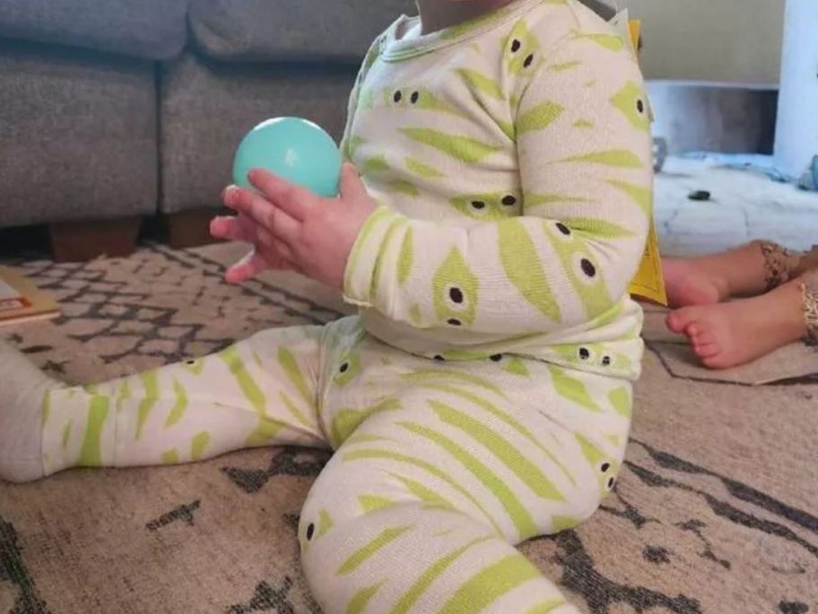 toddler wearing halloween pajamas while sitting on rug and holding ball