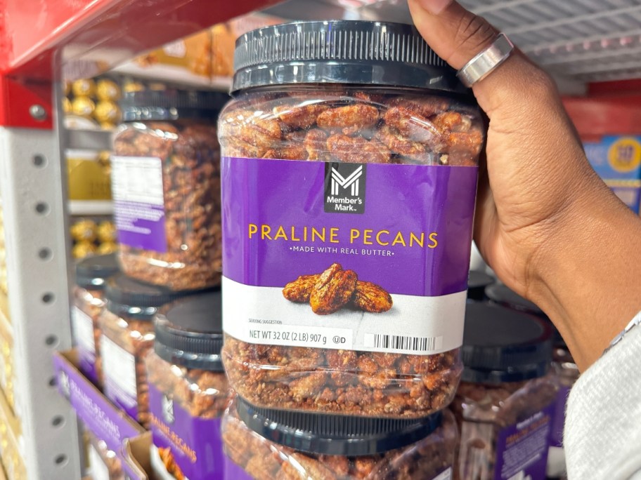 hand holding a large container of Member's Mark Praline Pecans 32oz Container with more on the shelf behind it