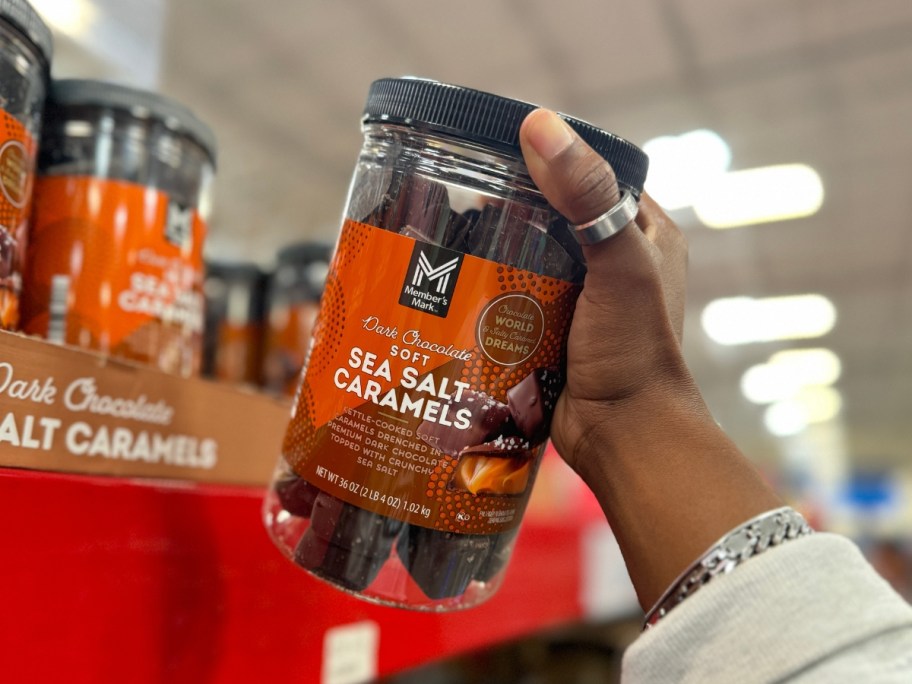 hand holding a jar with an orangish color label for Member's Mark Dark Chocolate Soft Sea Salt Caramels, more on a shelf behind it