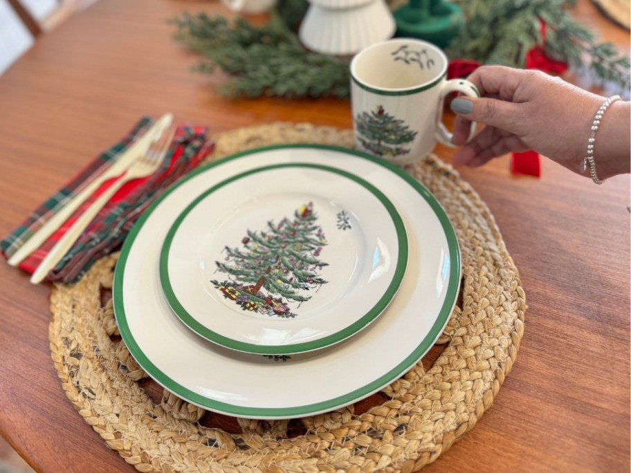 hand reaching for a cream color mug with a Christmas tree print on it on a table setting with a jute woven placemat and matching cream plate and salad plate with the same Christmas tree design