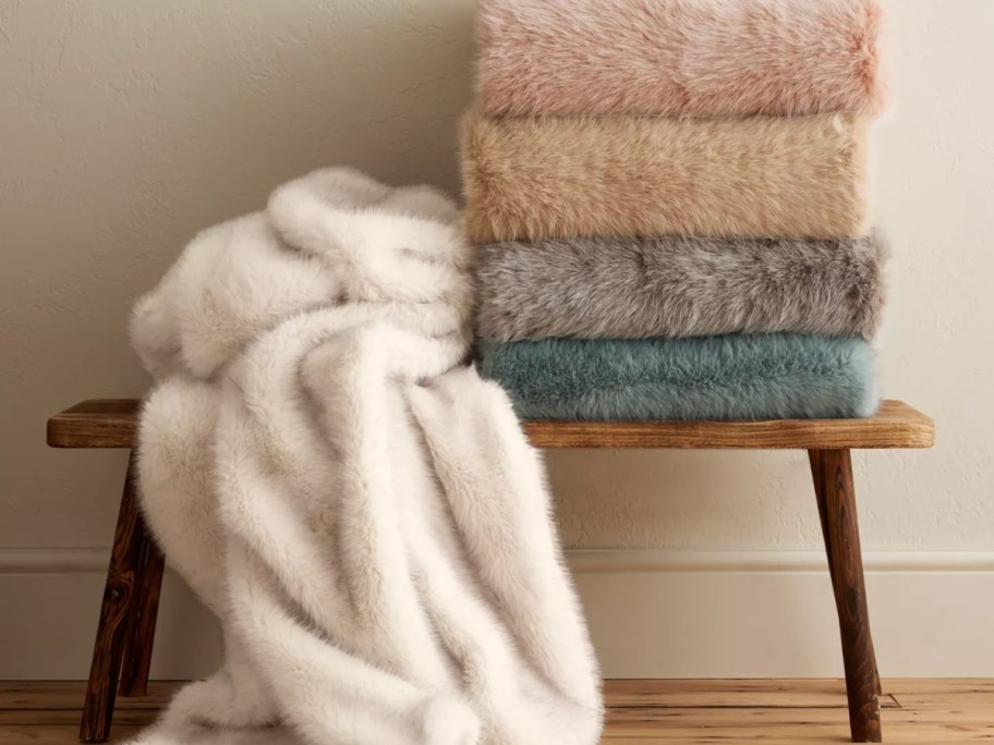 a cream faux fur blanket draped and falling off a bench, more of the same blanket in other colors stacked on the bench next to it