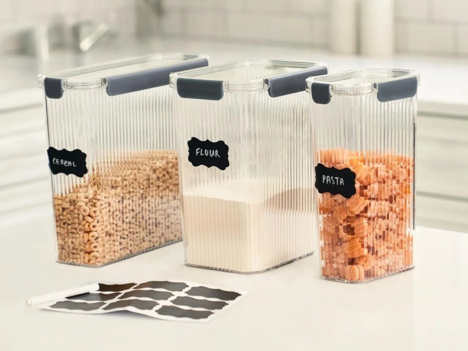 a set of 3 tall clear ribbed pantry food storage containers with snap lock lids and dry goods in them, black labels and a chalk pen are in front of them