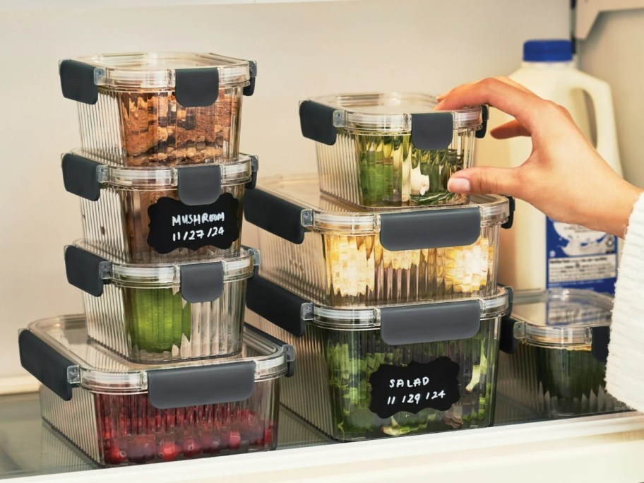 a set of clear ribbed food storage containers with snap lock lids with food in them in a fridge, they have black labels on them, a hand is reaching for 1