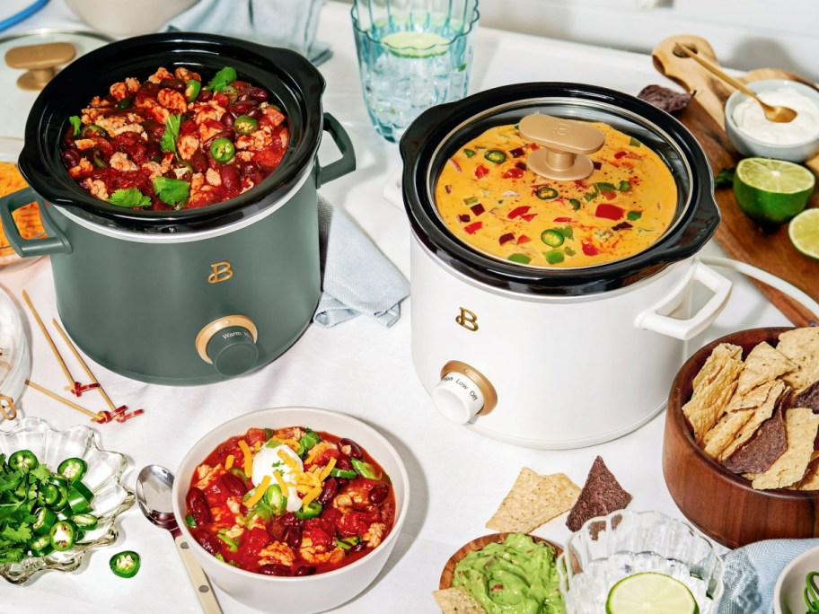 green and white Beautiful by Drew Barrymore 2-Quart Slow Cookers full of chili and queso dip and surrounded by a bowl of tortilla chips and other fixings.