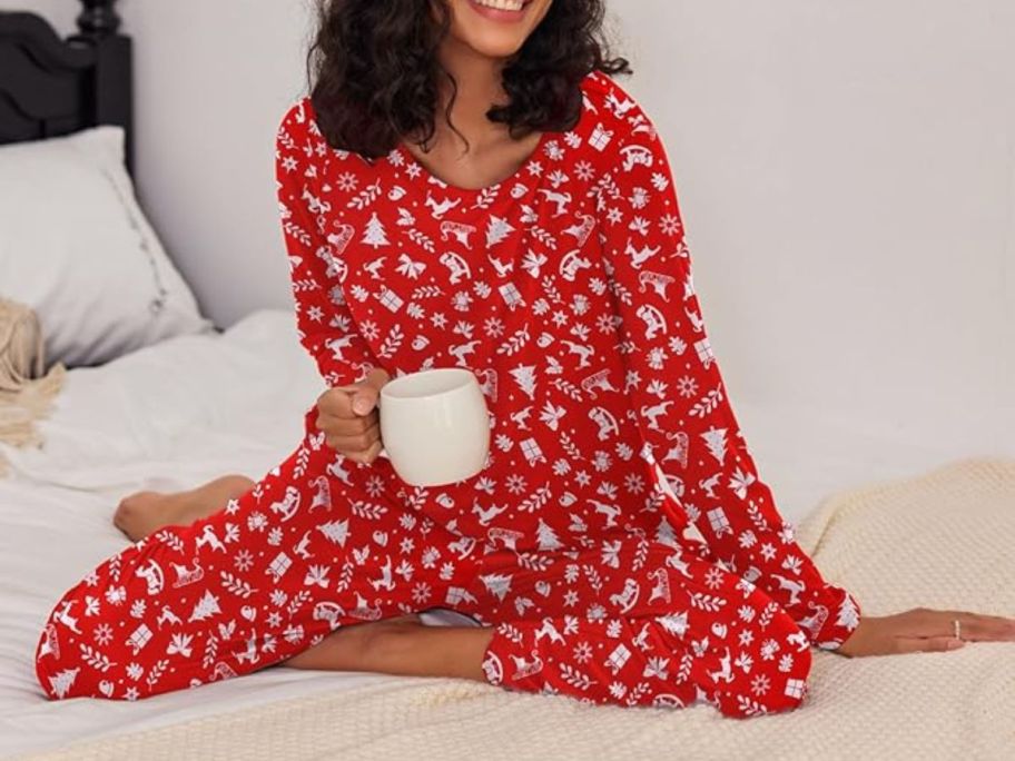 A woman sitting on a bed with a hot beverage wearing red Christmas pajamas 