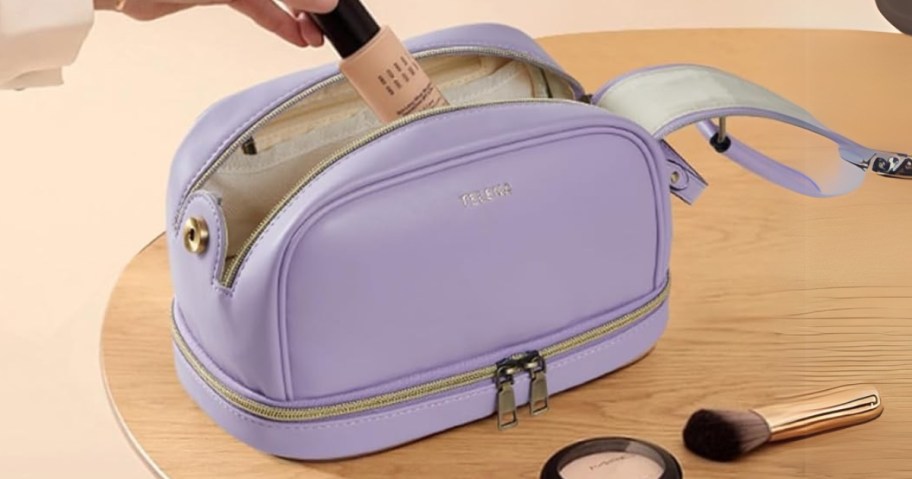 person putting a makeup bottle into a light purple makeup bag, more makeup on the table beside it