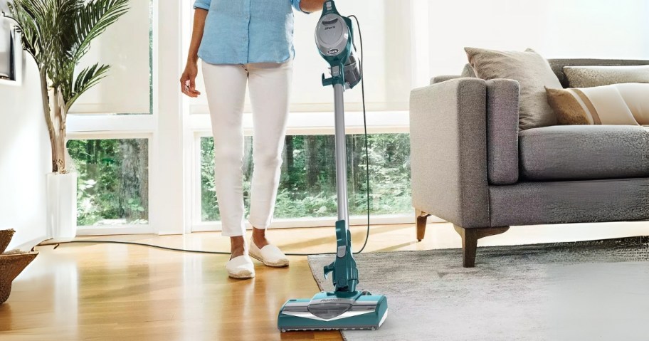 woman using a green corded stick Shark vacuum to vacuum the floor in a living room