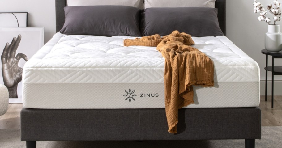 a white Zinus brand mattress on a grey platform bed frame with grey pillows and a goldish color throw blanket