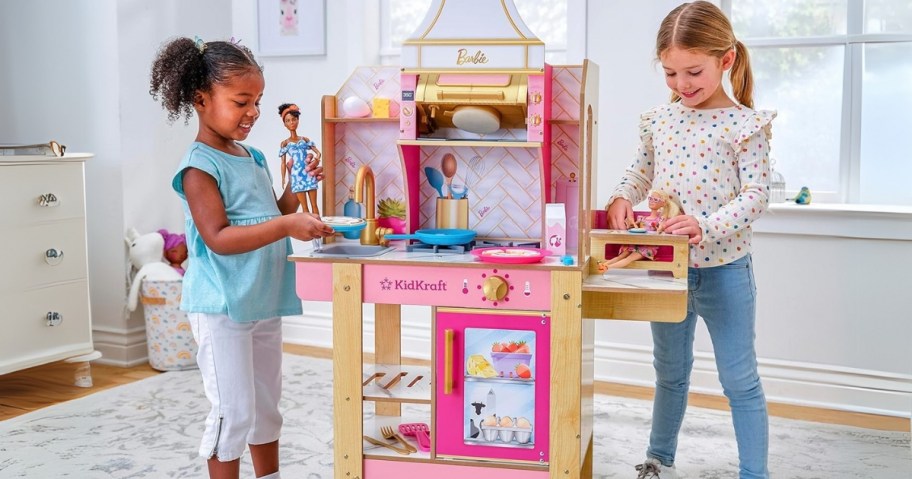 2 little girls playing with Barbies and a a kid's wooden toy kitchen in pink with Barbie logo and design features