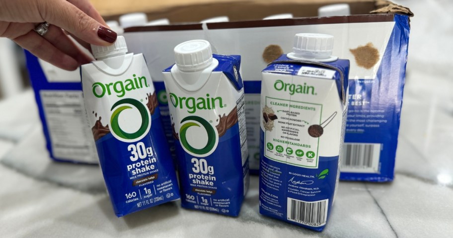 a hand reaching for a ready to drink Orgain shake bottle, 2 more beside it and box that a pack of 12 comes in behind it