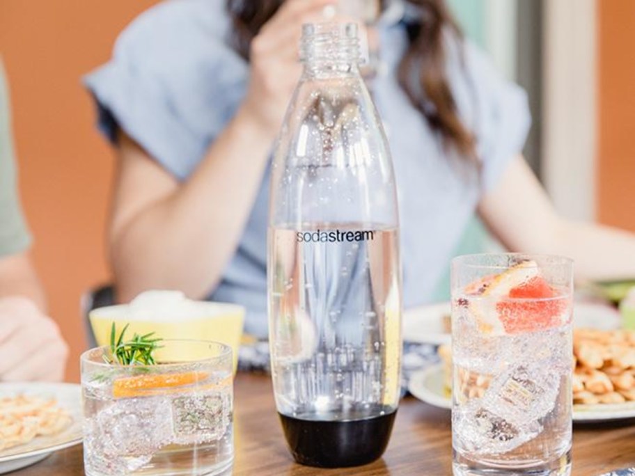 a clear an black SodaStream 1L bottle with carbonated water in it, 2 glasses with water and fruit beside it, woman sitting behind it.