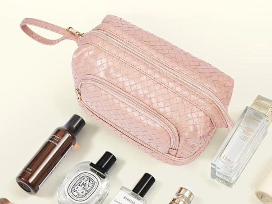 a pink faux leather makeup bag with several compartments, beauty items laying in front of it