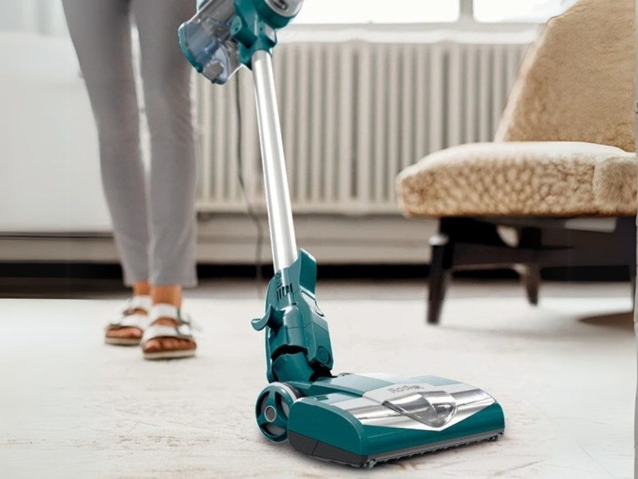 woman using a green corded stick Shark vacuum to vacuum the floor in a living room