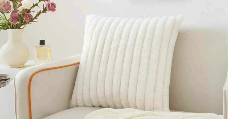 a cream color ribbed faux fur pillow on a tan sofa next to a side table with a vase of flowers on it